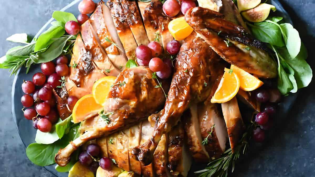 A beautifully roasted turkey garnished with fresh herbs, orange slices, and clusters of red grapes. The turkey is sliced on a platter, surrounded by leafy greens and figs, creating a vibrant, appetizing presentation.