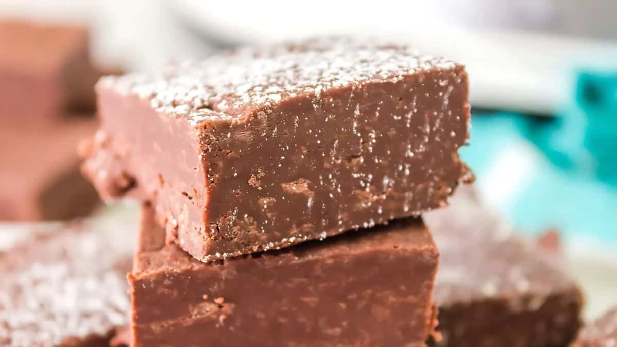 A close-up of stacked chocolate fudge squares dusted with powdered sugar. The texture looks rich and creamy, with a glossy finish.