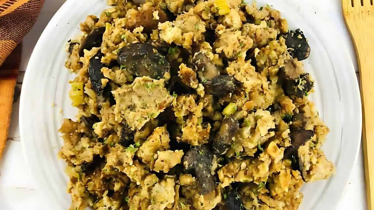 A white plate filled with a savory dish featuring crumbled bread pieces, sliced mushrooms, and chopped herbs. A wooden fork is partially visible on the side. The dish has a rustic and hearty appearance.