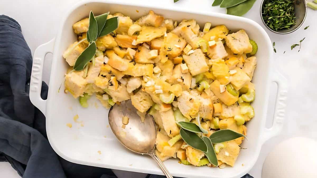 A white baking dish filled with chunky bread stuffing, garnished with fresh sage leaves and thyme. Diced celery is visible among the bread pieces. A silver spoon rests inside the dish, and a small bowl of chopped herbs is nearby.
