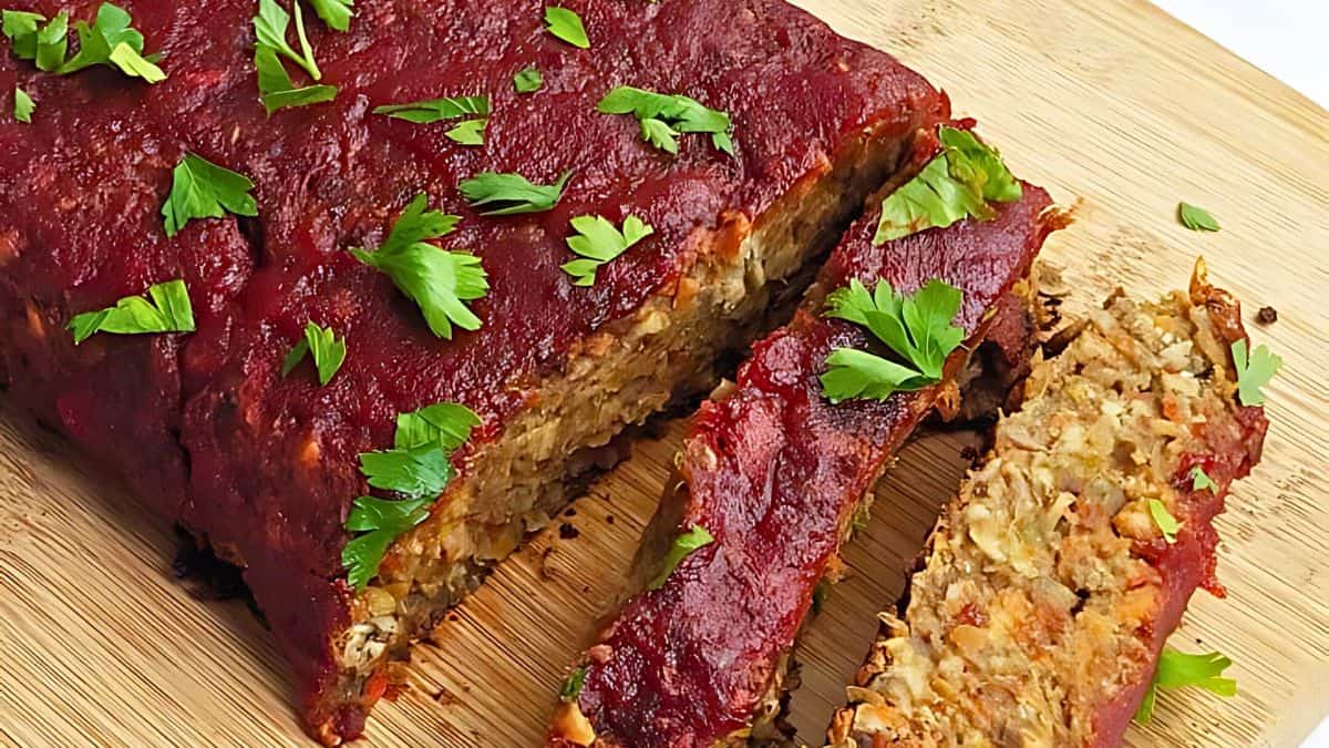 A sliced meatloaf topped with red sauce is garnished with fresh parsley. It rests on a wooden cutting board with pieces cut from the loaf, revealing a savory and textured filling.