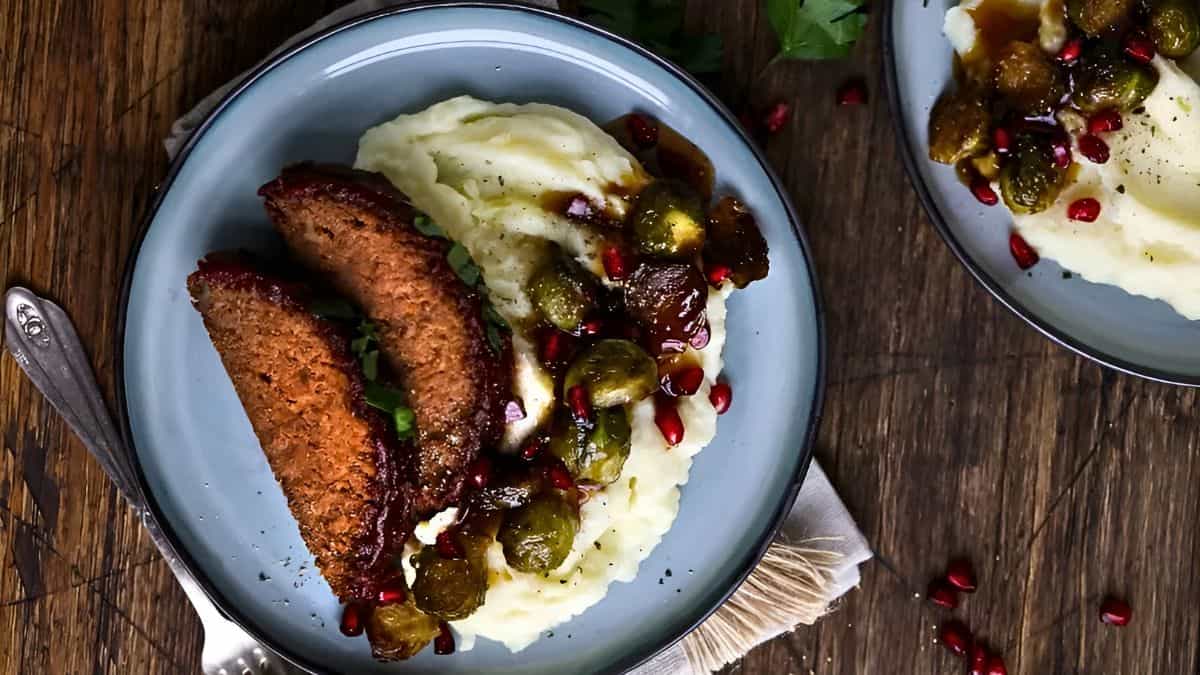 A plate of mashed potatoes topped with roasted Brussels sprouts and pomegranate seeds, accompanied by two slices of seasoned meatloaf. The dish is served on a rustic wooden table.