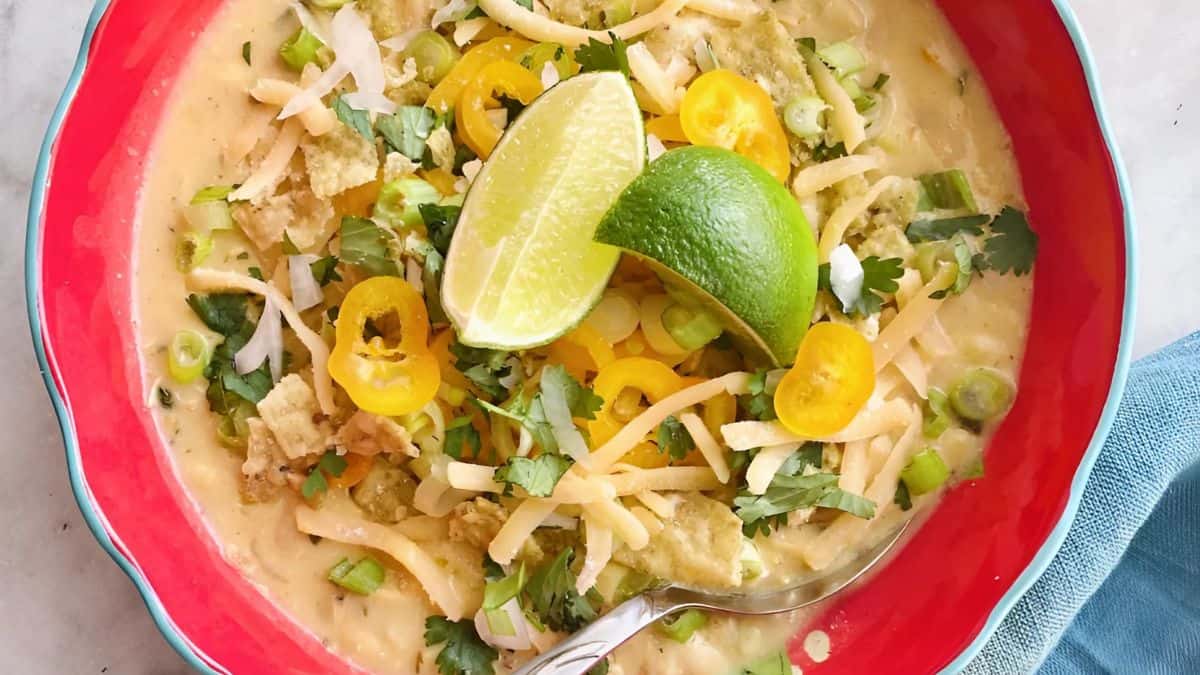 A colorful bowl of creamy soup garnished with shredded cheese, sliced yellow peppers, chopped green onions, cilantro, crushed tortilla chips, and a lime wedge, served in a vibrant red bowl with a silver spoon on the side.