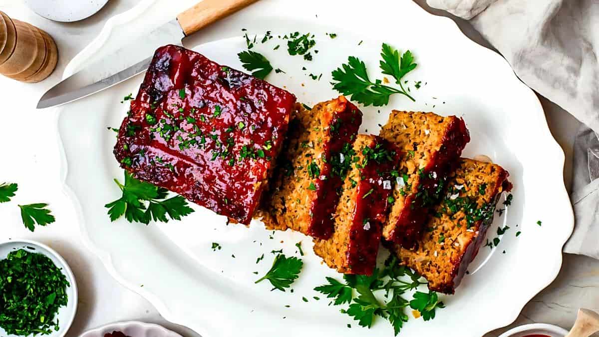 Sliced meatloaf with a glossy glaze is served on a white platter, garnished with fresh parsley. There are chopped herbs in small bowls nearby, and a knife rests beside the meatloaf.