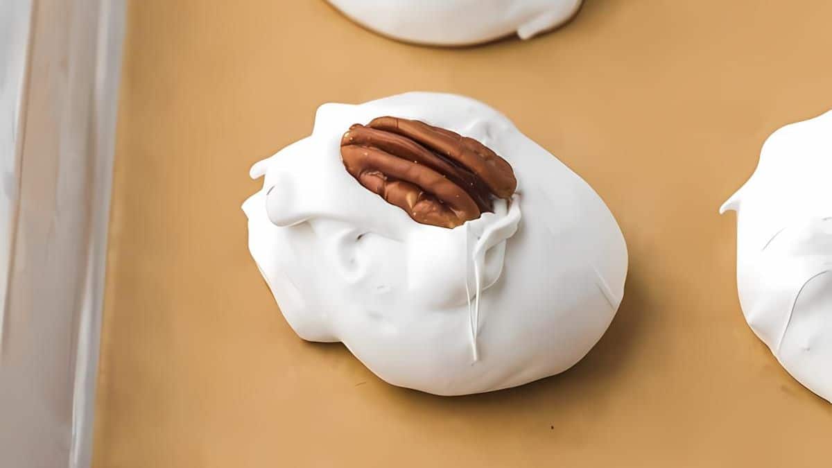 A close-up of a meringue cookie topped with a pecan on a brown parchment-lined baking sheet. The meringue is glossy and white, with a slightly uneven texture.