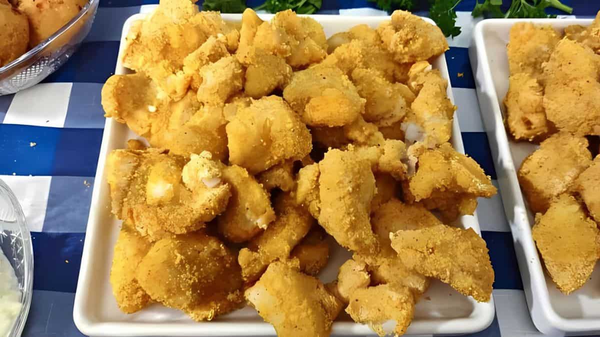 A plate filled with golden-brown, breaded and fried fish nuggets, placed on a blue and white checkered tablecloth. Other dishes and greenery are partially visible in the background.