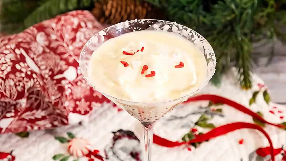 A festive cocktail in a martini glass, garnished with whipped cream and small red heart sprinkles. The background features holiday-themed decorations with greenery, a red patterned cloth, and ribbons.