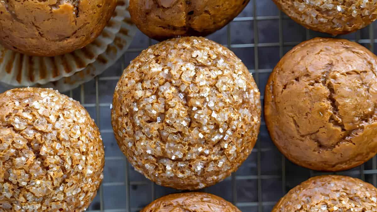 A close-up of several golden-brown muffins resting on a cooling rack. The muffins are topped with a generous layer of sparkling sugar crystals, giving them a textured appearance. One muffin is partially unwrapped from its paper liner.