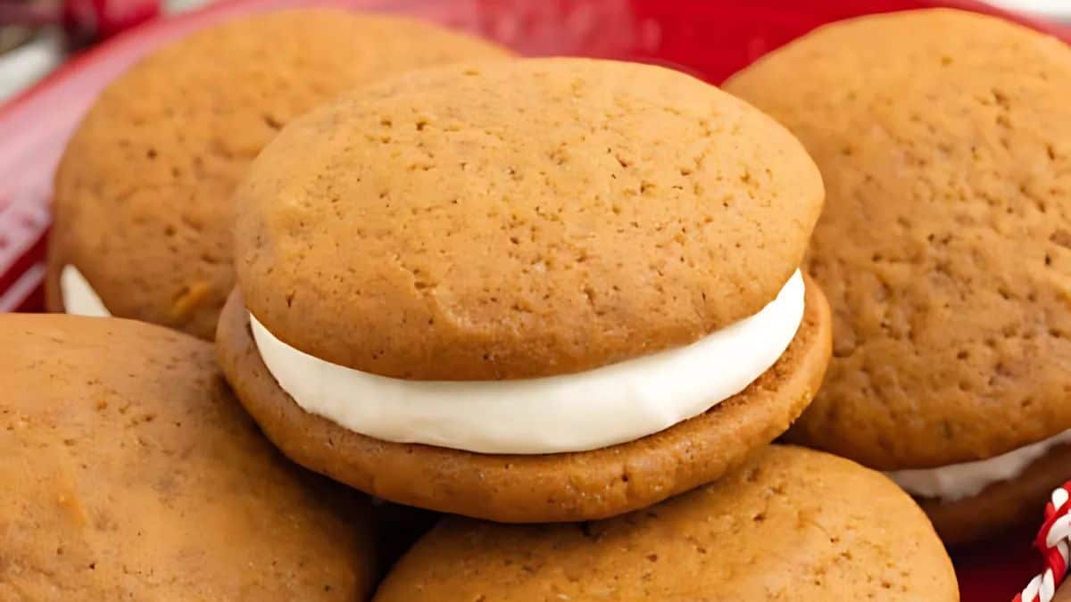 Close-up of several gingerbread whoopie pies with cream filling, stacked on a red plate. The cookies are golden brown with a smooth texture.