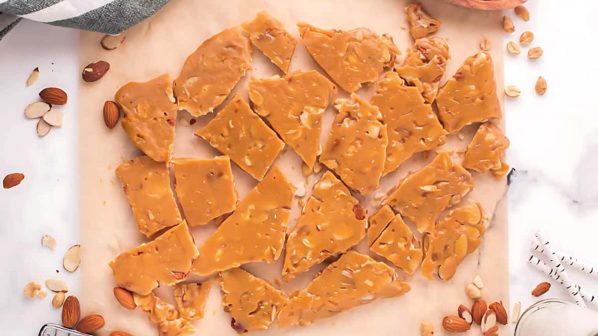 Broken pieces of homemade peanut brittle rest on parchment paper. Surrounded by scattered almonds and peanuts, the golden-brown candy has a glossy appearance, showcasing visible nut pieces throughout. A striped cloth is visible in the corner.
