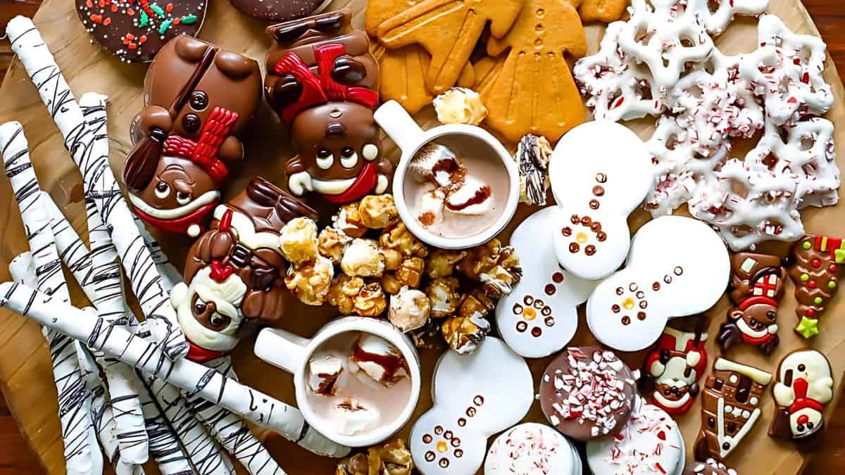 An assortment of holiday-themed chocolates and treats on a table. Includes chocolate-dipped pretzels, snowman and reindeer-shaped chocolates, caramel corn, cookies, and two mugs of hot chocolate topped with marshmallows.