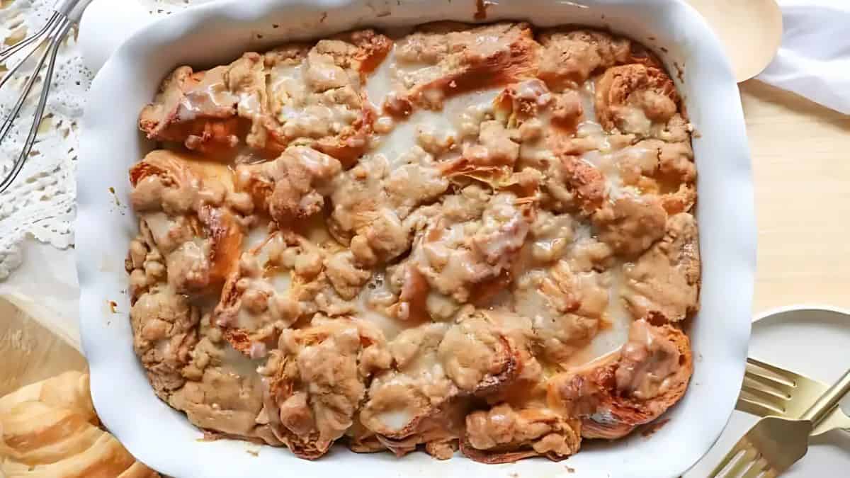 A baked dish of cinnamon roll casserole with a golden-brown crust and drizzled icing, served in a white rectangular baking dish. A fork and a plate are nearby on a wooden table.