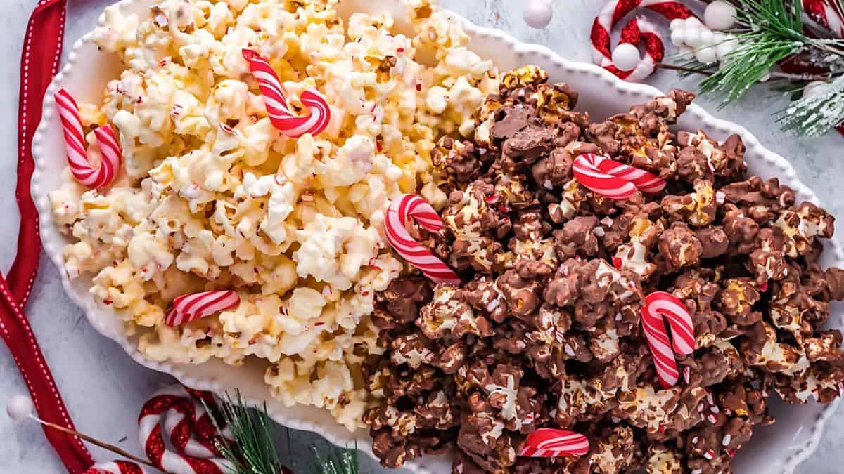 A festive platter featuring white popcorn and chocolate-covered popcorn, garnished with small candy canes. Surrounding the dish are holiday decorations, including greenery and red ribbon.