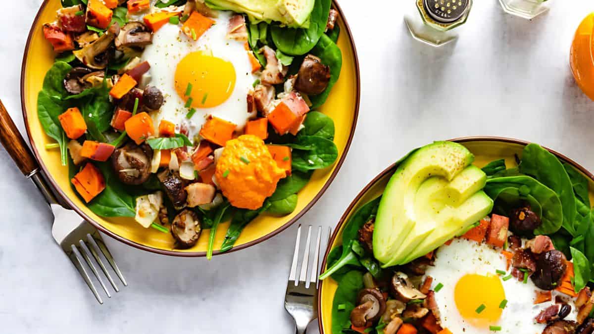 Two bowls filled with vibrant breakfast salads, each featuring a sunny-side-up egg atop spinach, mushrooms, diced sweet potatoes, avocado slices, and chopped vegetables. Forks are placed beside the bowls on a white background.