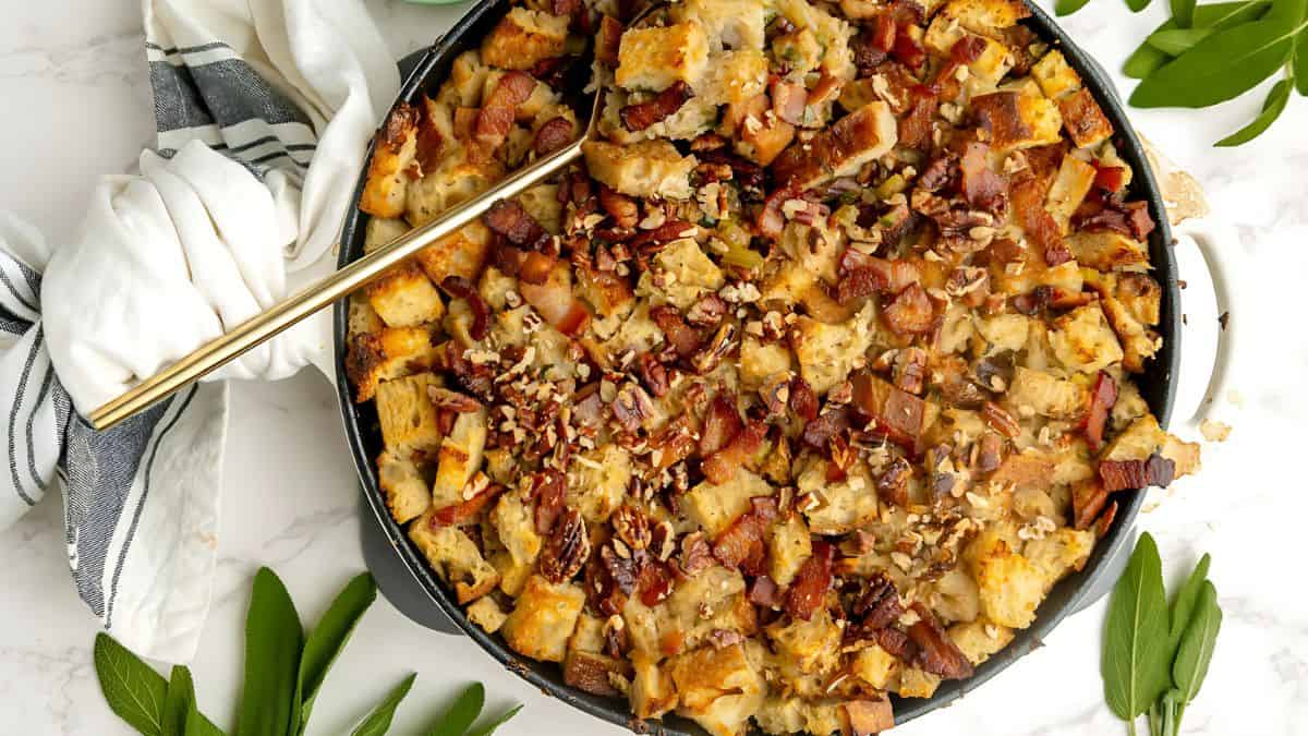 A top-view of a skillet filled with stuffing. The dish features chunks of bread, bacon, pecans, and herbs. A striped napkin and large spoon lie on the side. Fresh green leaves are scattered around the skillet on the white surface.