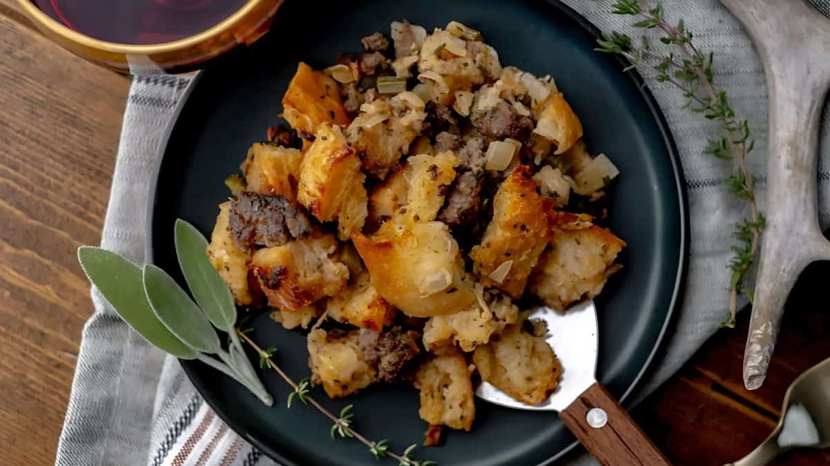 A plate of savory stuffing with chunks of bread, herbs, onions, and meat, garnished with fresh sage and thyme. The dish rests on a striped cloth with a wooden-handled serving spoon nearby. A glass of red wine is visible in the background.