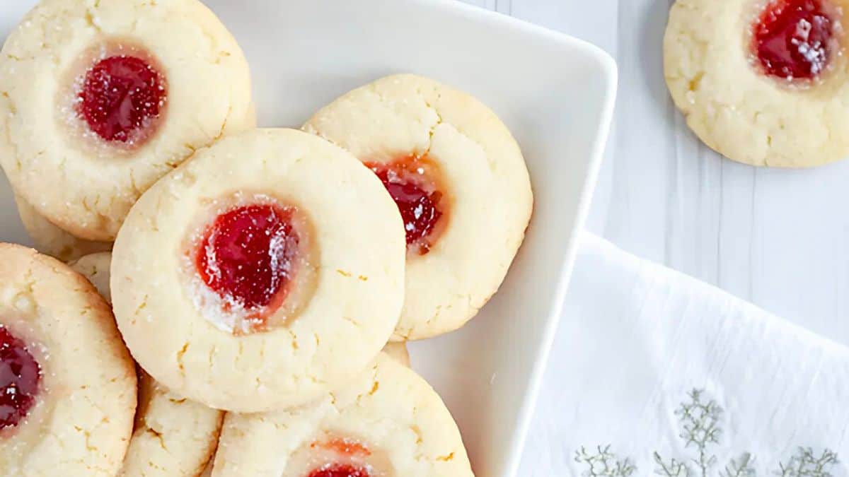 A white plate with several thumbprint cookies filled with red jam, arranged neatly. One cookie is placed on a white tablecloth nearby. The cookies are light golden-brown with a sugary texture.