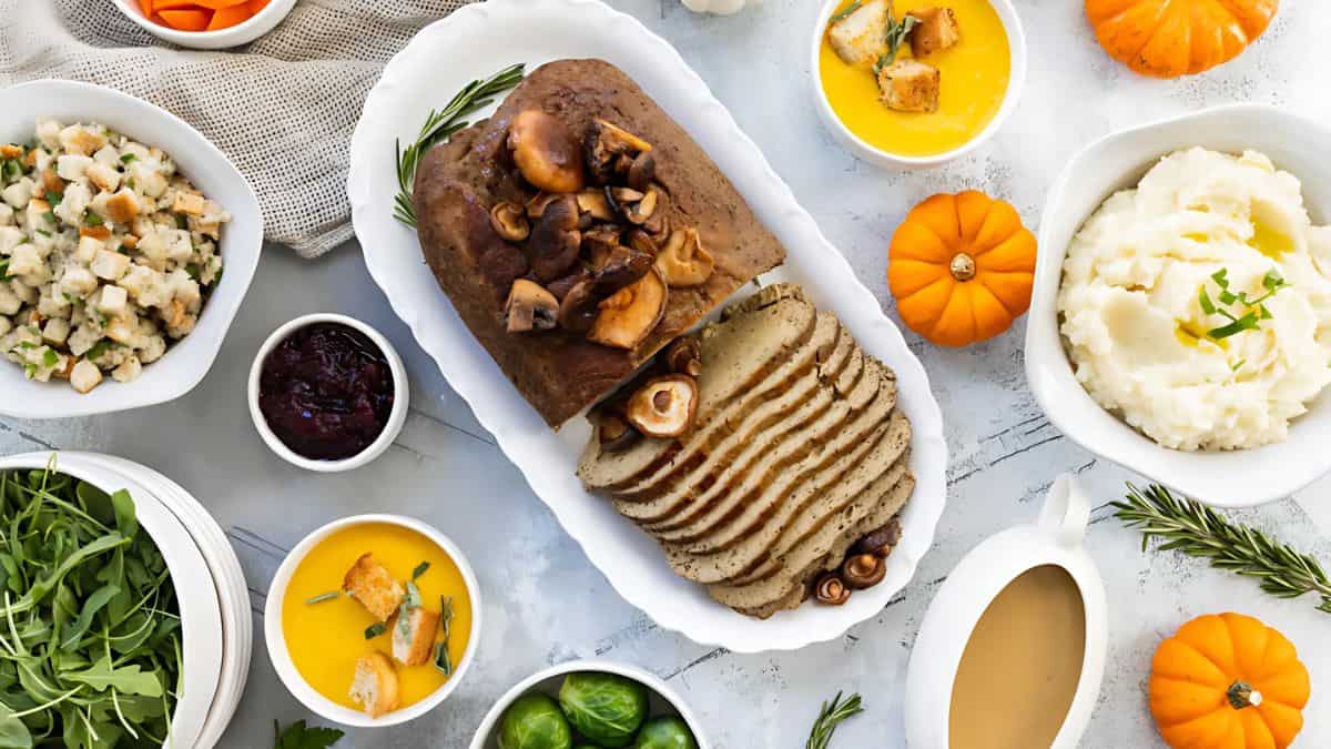 A Thanksgiving meal featuring a loaf with mushrooms on a platter, surrounded by mashed potatoes, stuffing, cranberry sauce, gravy, small pumpkins, and bowls of soup, salad, and Brussels sprouts, all arranged on a white table.