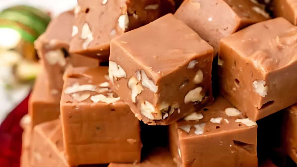 A close-up of a pile of square, creamy fudge pieces with visible nut chunks throughout. The fudge is arranged neatly, showcasing its smooth texture and rich brown color.