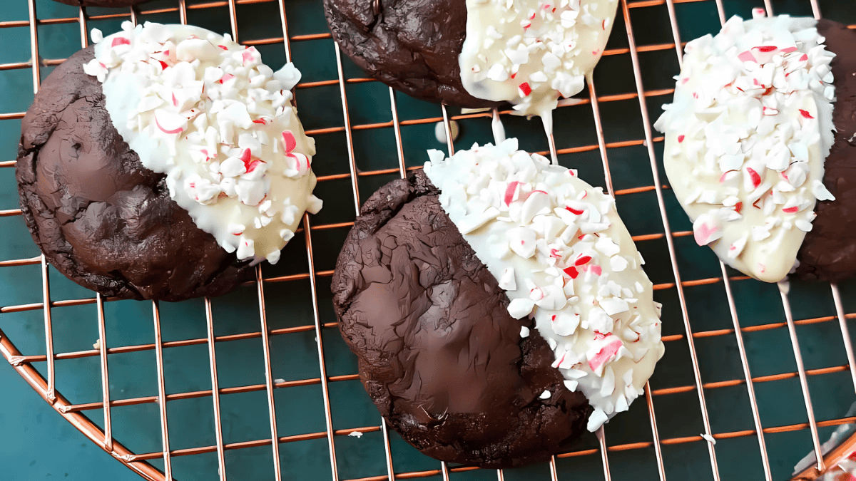 Four chocolate cookies, half-dipped in white chocolate and topped with crushed peppermint, rest on a wire cooling rack.