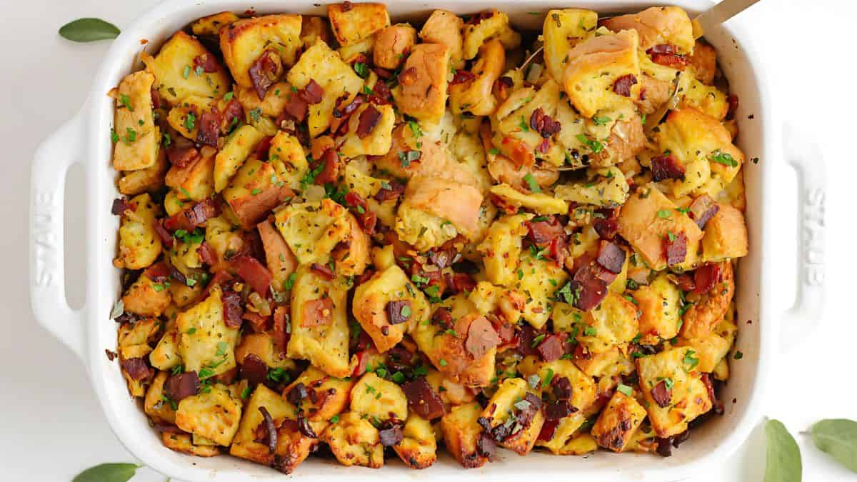 A white casserole dish filled with golden-brown stuffing, featuring chunks of bread, crispy bacon, and sprinkled with chopped herbs.