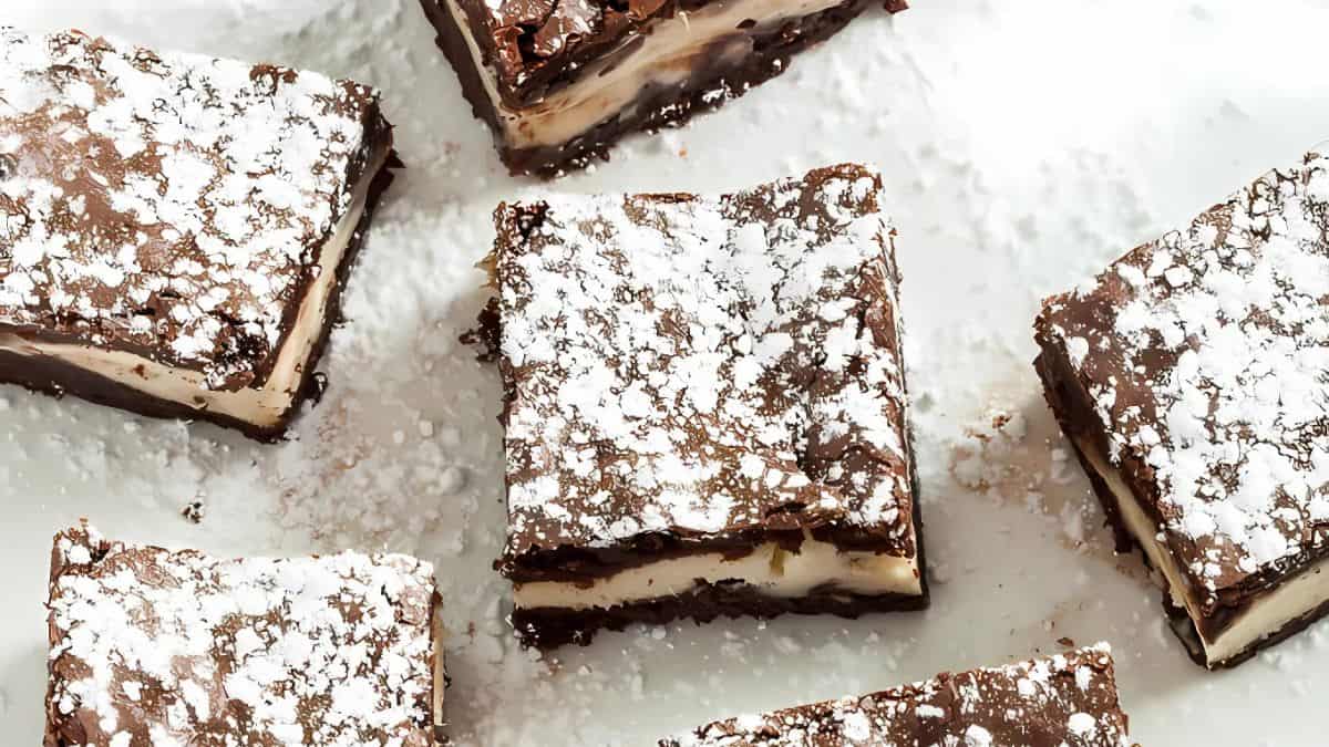 Five chocolate brownies are topped with powdered sugar, arranged on a white surface. Some have a visible cream layer, revealing a rich and decadent texture.