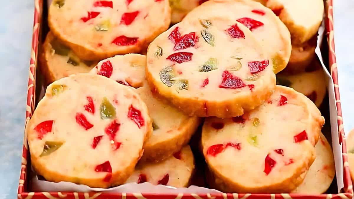 A close-up of a patterned box filled with round cookies studded with red and green candied fruits. The cookies are light brown with a slightly crumbly texture, arranged neatly inside the box.