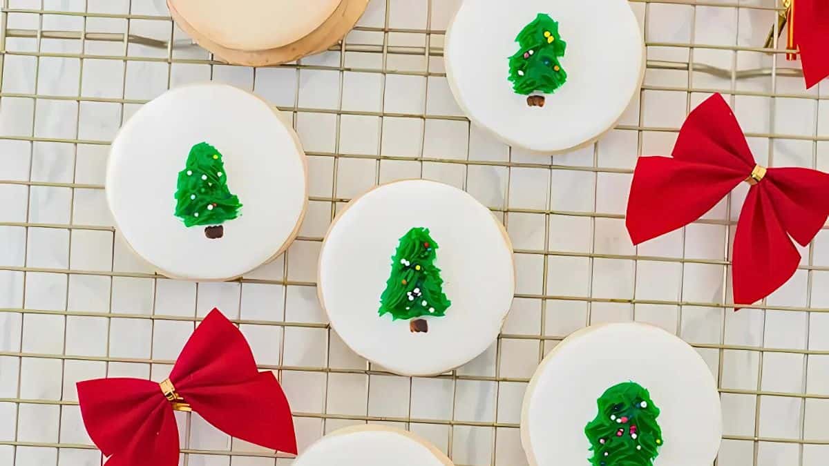 Round cookies with white icing are decorated with small green Christmas trees, topped with sprinkles. Red bows are scattered around the cookies, all placed on a wire cooling rack.