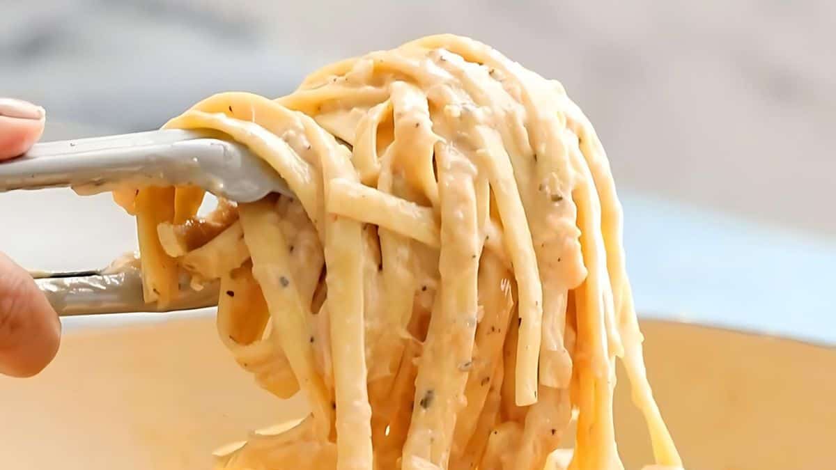 Close-up of creamy pasta being lifted with tongs above a bowl. The pasta is coated in a rich, textured sauce with herbs visible, creating an appetizing and savory appearance.