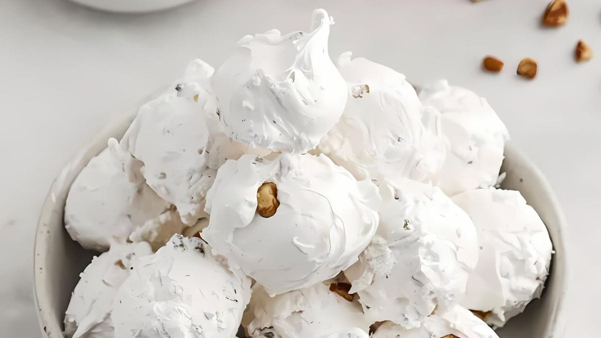 A bowl filled with fluffy white divinity candy, with small nut pieces visible throughout. A few nuts are scattered on the white surface beside the bowl.