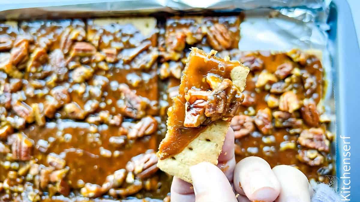 A hand holding a pecan praline cracker over a tray filled with more crackers, topped with caramel and pecans, set on a silver baking sheet in the background.