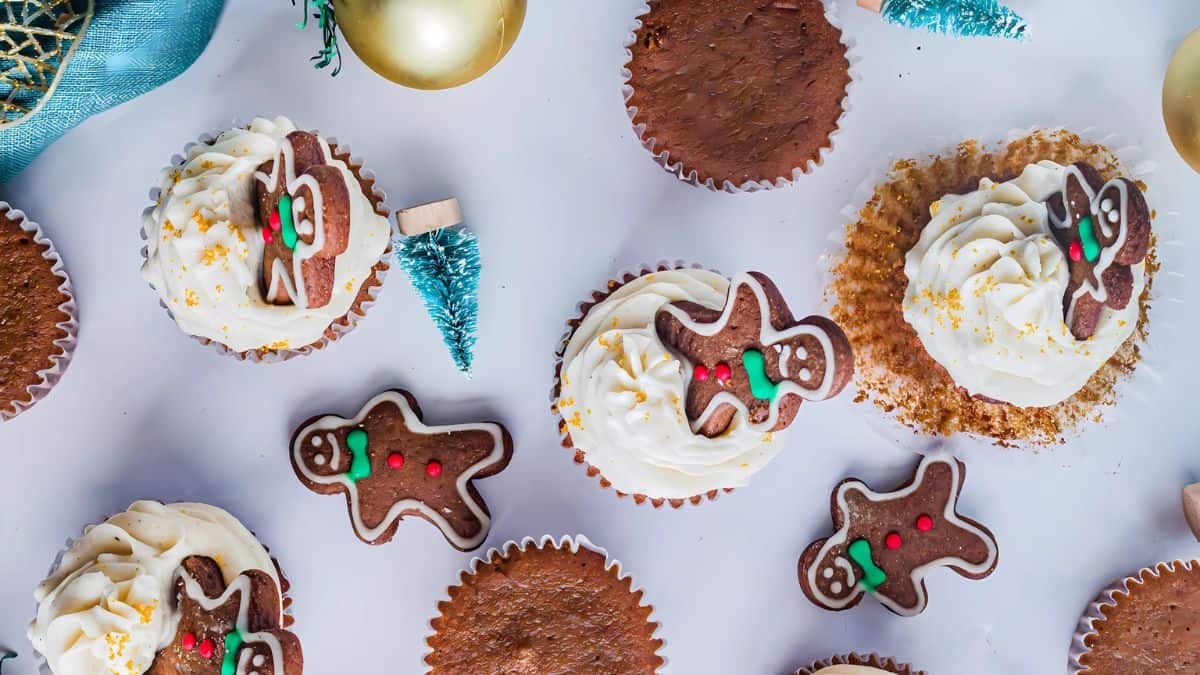 Top view of festive cupcakes decorated with gingerbread men and cream, surrounded by small blue Christmas trees and golden ornaments on a white background.