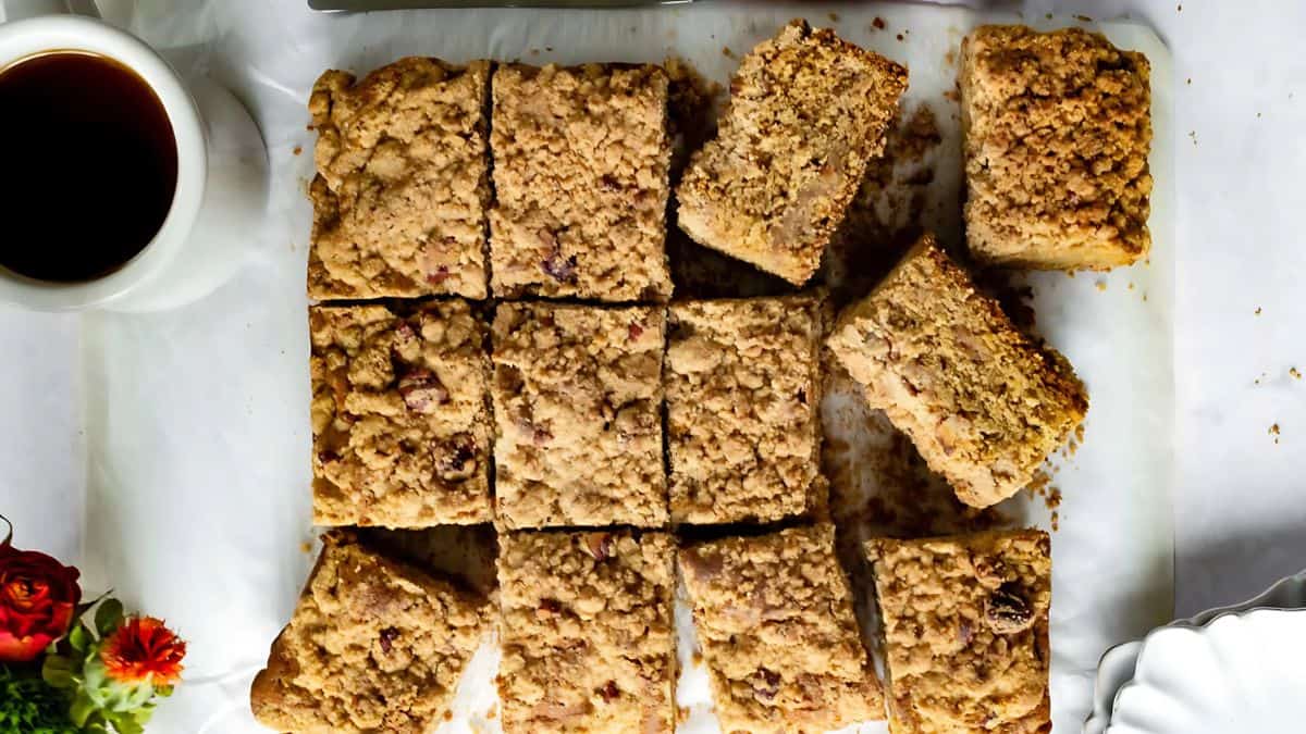 Thirteen pieces of crumbly coffee cake with a streusel topping are arranged on a parchment-lined surface. A cup of black coffee and a small red flower are placed nearby for a cozy presentation.