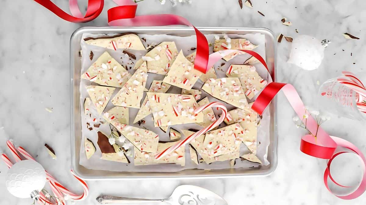 A tray of peppermint bark pieces decorated with crushed candy canes and red ribbon on a marble surface. Surrounded by candy canes, white ornaments, and flowers.