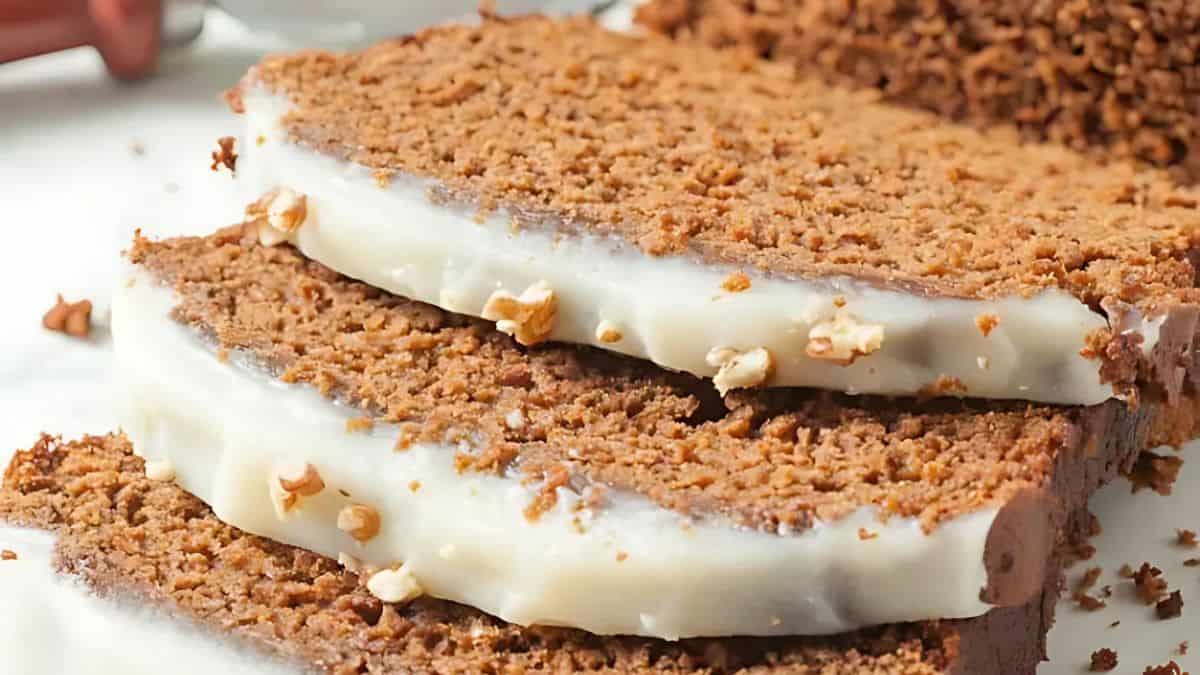 Two slices of spiced loaf cake with a creamy white frosting, topped with small nuts, are stacked on a plate. Crumbs are scattered around, and more cake is visible in the background.