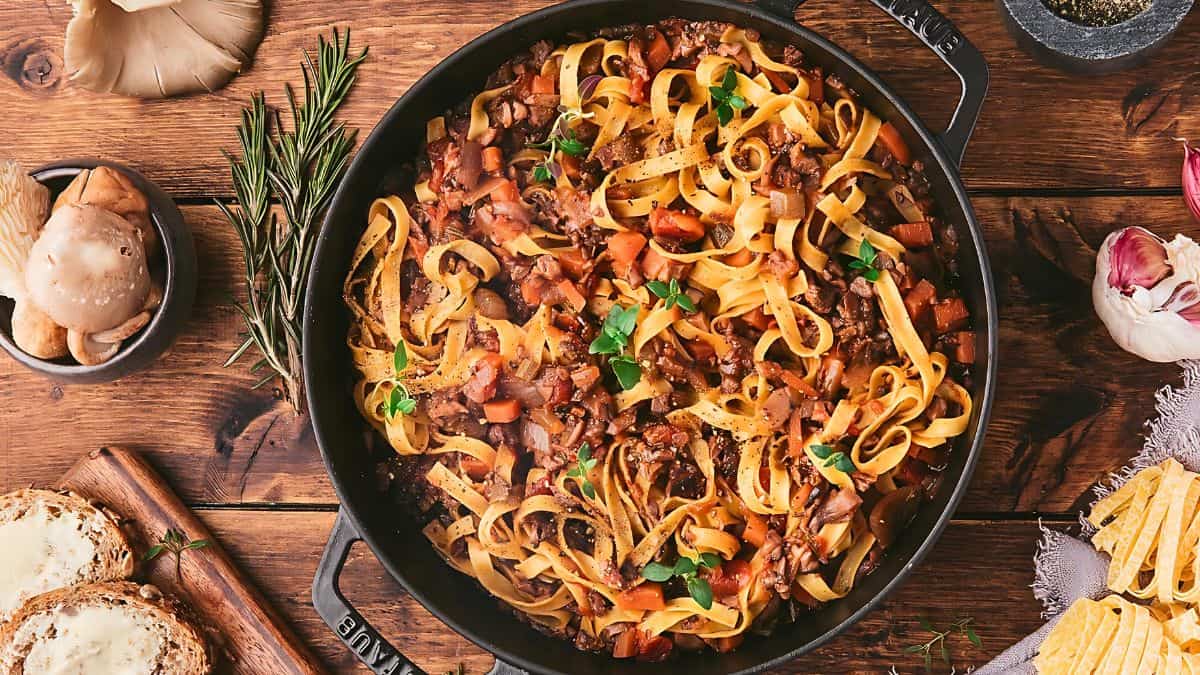A pan of tagliatelle pasta mixed with a hearty bolognese sauce, garnished with herbs. Surrounding the pan are slices of bread, garlic cloves, rosemary sprigs, mushrooms, and a small bowl of pepper, all set on a rustic wooden table.