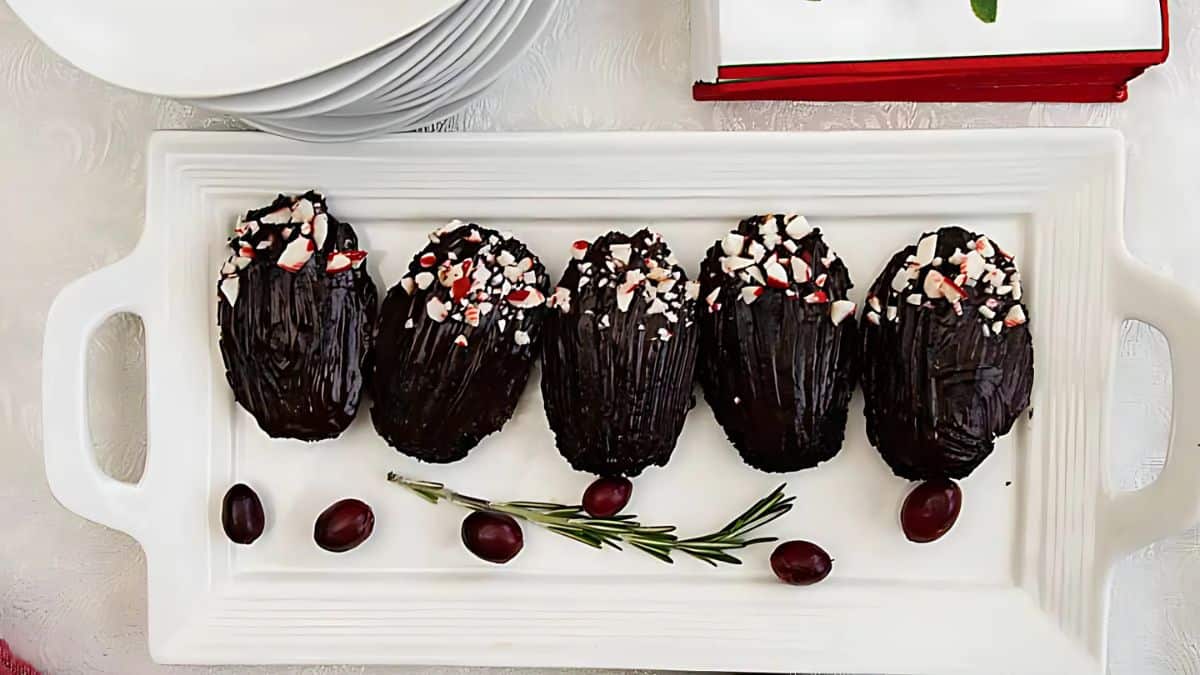 A white rectangular platter holds five dark chocolate treats topped with crushed peppermint pieces. Garnished with rosemary sprigs and a few cherries on a light table.
