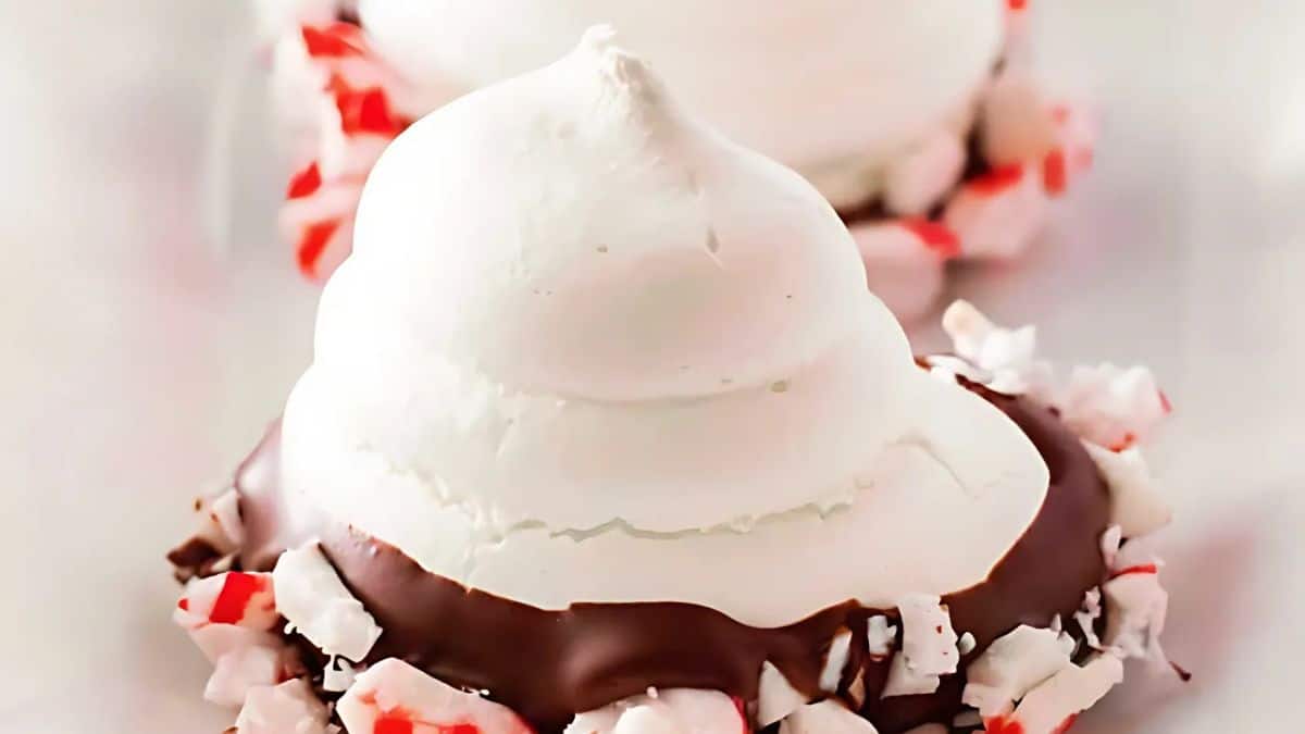 Close-up of a dessert featuring a dollop of white meringue on top of a chocolate-covered base. The edges are coated with crushed peppermint pieces, adding a festive touch. The background is blurred, focusing on the intricate details of the treat.