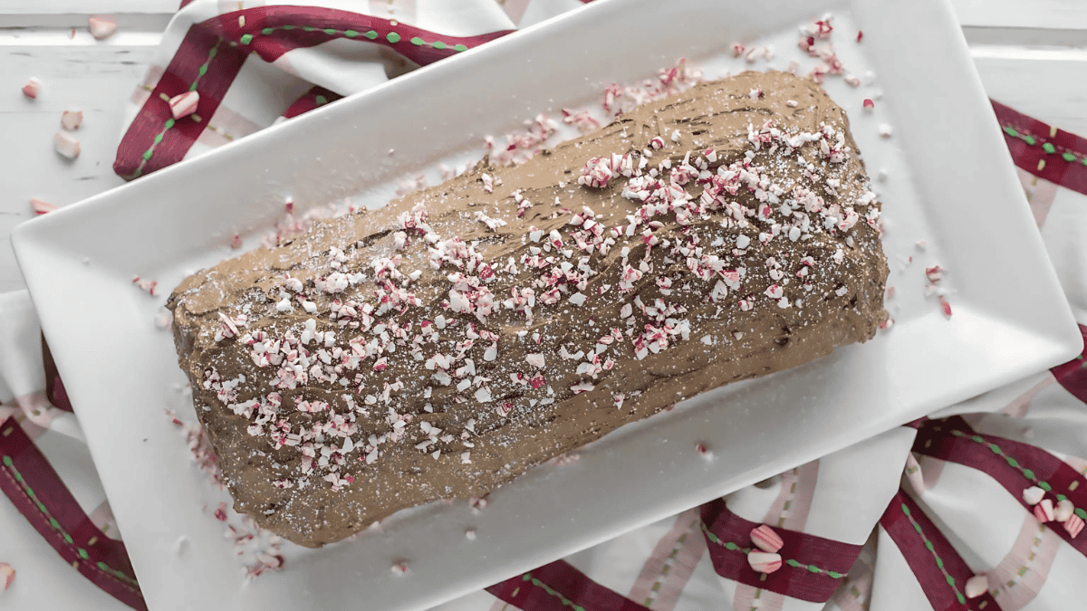 A chocolate yule log cake on a rectangular white platter, topped with crushed peppermint candies. The platter is placed on a white surface with a red and white striped cloth underneath.