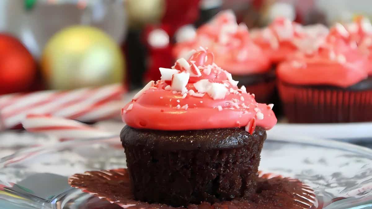 A chocolate cupcake with bright pink frosting and white sprinkles is unwrapped on a plate. In the background, similar cupcakes are arranged with festive decorations like red ornaments and candy canes, creating a holiday atmosphere.