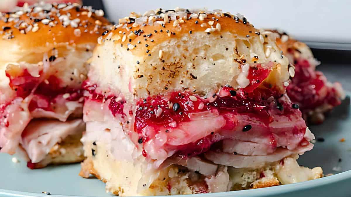 Close-up of two turkey sliders on a plate, layered with cranberry sauce and melted cheese. The top bun is sprinkled with sesame and poppy seeds, offering a festive and colorful presentation.