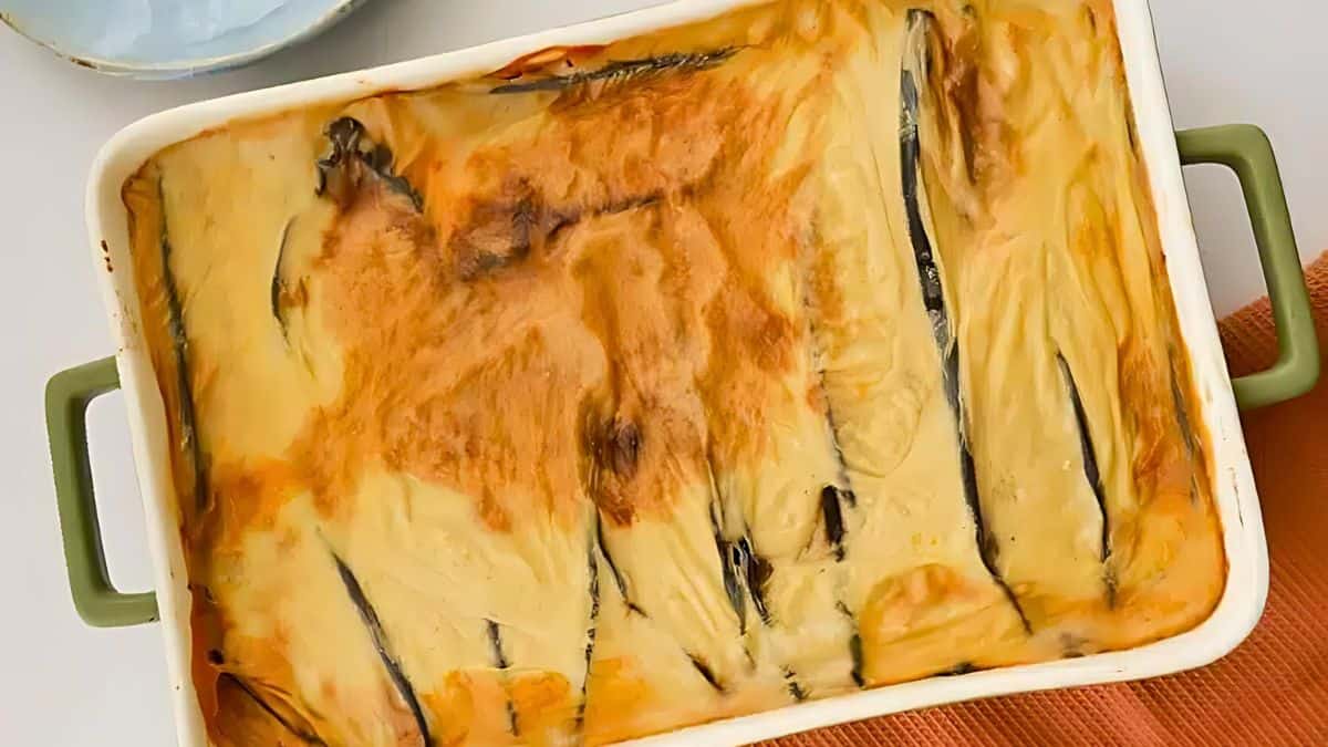 A baked moussaka dish in a rectangular green-handled pan. The top is golden brown, with visible layers of eggplant underneath. A textured cloth is partially visible in the bottom corner.