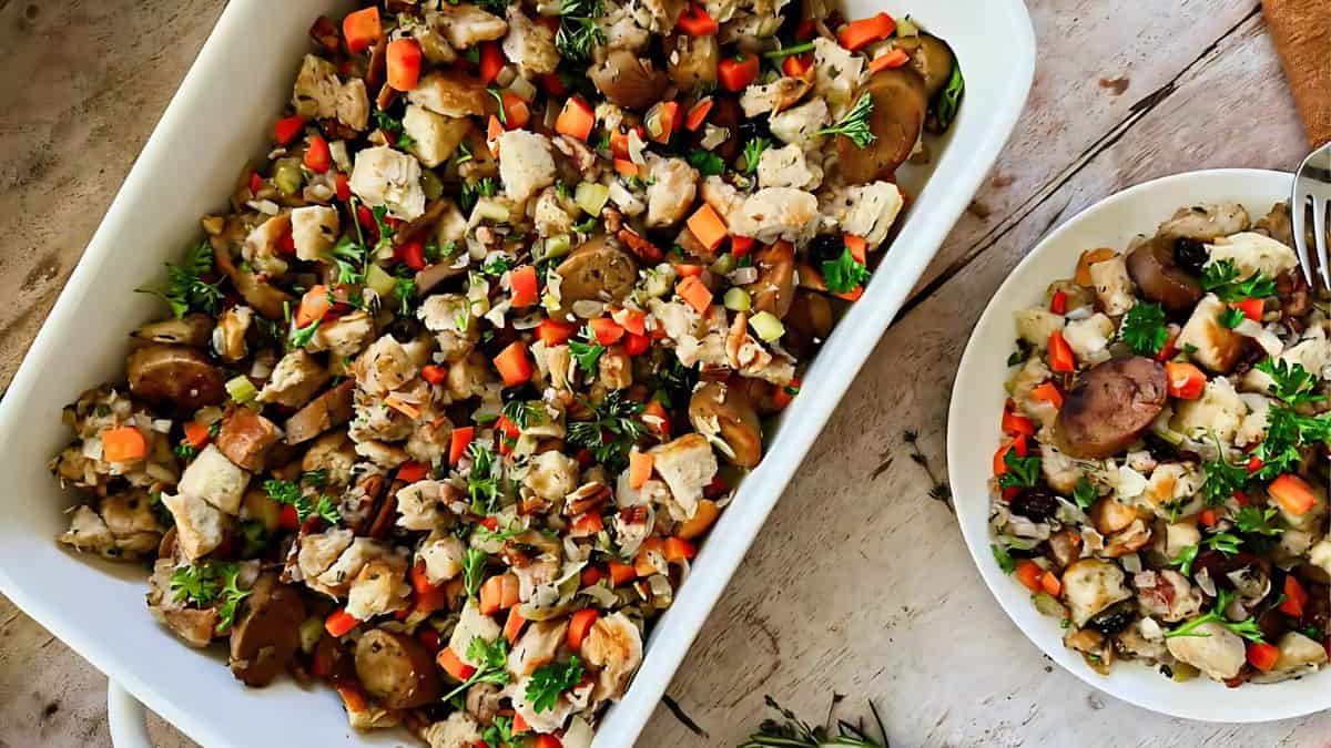 A casserole dish filled with a savory stuffing mix of diced bread, vegetables, mushrooms, and herbs, paired with a serving on a side plate. The dish is garnished with fresh green herbs, set on a rustic wooden table.
