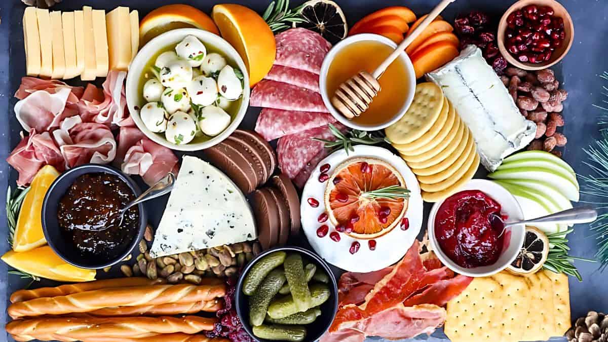 A vibrant charcuterie board filled with assorted cheeses, meats, crackers, sliced fruits, pickles, nuts, and preserves. There's an arrangement of mozzarella balls, blue cheese, honey, and garnished with fresh herbs and pomegranate seeds.