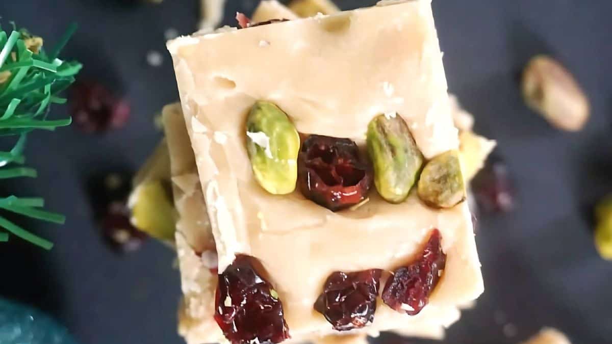 A close-up of a slice of white chocolate fudge garnished with pistachios and dried cranberries, set on a dark surface. A small green decoration is partially visible at the edge.