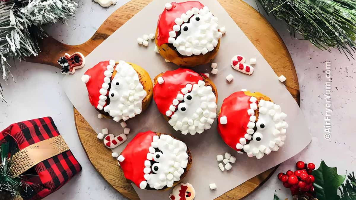 Five cupcakes decorated as Santa faces with red hats and white beards made of frosting and marshmallows. They are placed on a wooden board surrounded by a small gift and festive decorations.