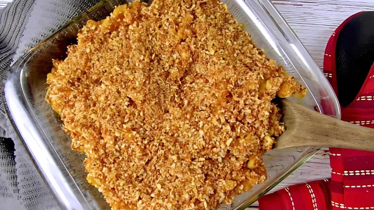 A glass baking dish filled with golden-brown baked macaroni and cheese topped with crunchy breadcrumbs. A wooden spoon is resting in the dish, and a red and black oven mitt is visible beside it.