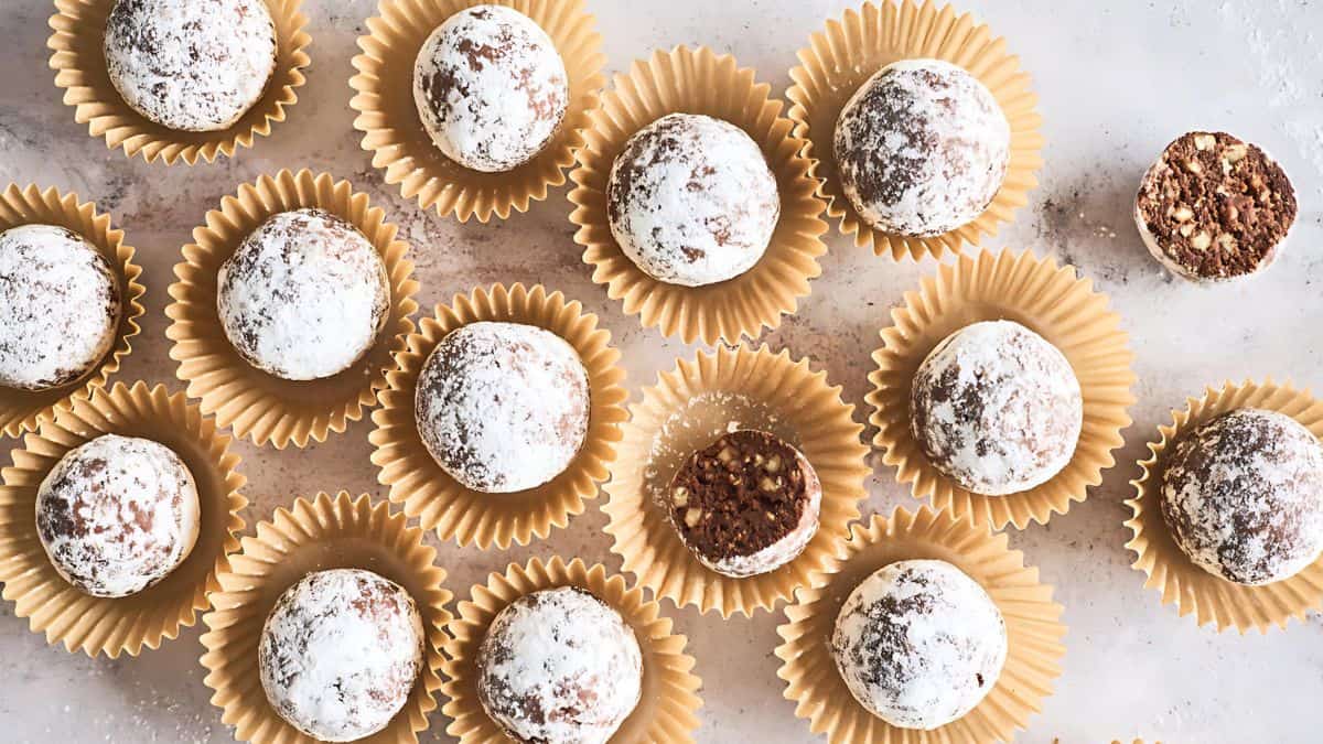 A collection of chocolate truffles dusted with powdered sugar, arranged in brown paper cups on a light surface. One truffle is cut in half, revealing a nutty filling.