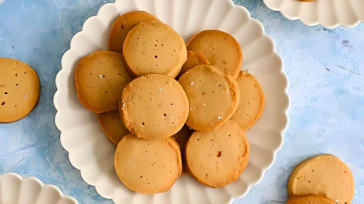 A white scalloped plate filled with round, golden-brown cookies sprinkled with salt. The background is a light blue surface with additional plates of cookies partially visible around the main plate.