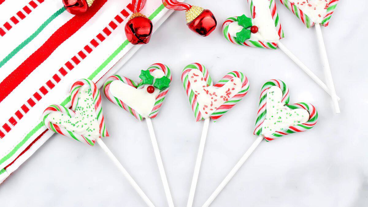 Heart-shaped candy canes with white chocolate and festive sprinkles, decorated with green holly and a red berry, on lollipop sticks. They are arranged on a white surface near a red and white striped cloth and red jingle bells.